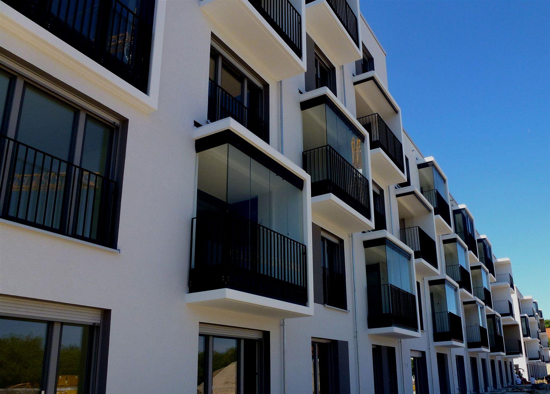 Das Bild zeigt eine moderne Wohnhausfassade mit vielen Glasbalkonen und klarer, geometrischer Architektur bei blauem Himmel.