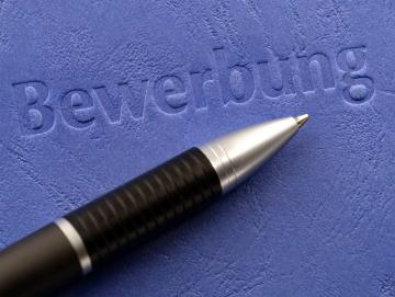 Das Bild zeigt einen Kugelschreiber, der auf einer blauen Bewerbungsmappe liegt. Auf der Mappe ist das Wort „Bewerbung“ geprägt.