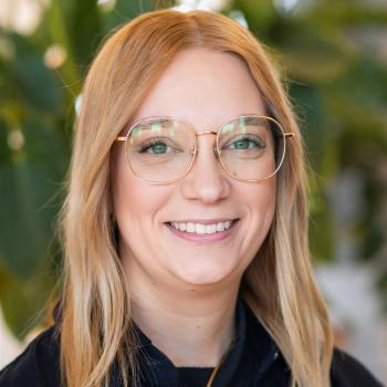 Das Bild zeigt eine Frau mit langen, blonden Haaren und runder Brille, im Hintergrund sind Pflanzen erkennbar.