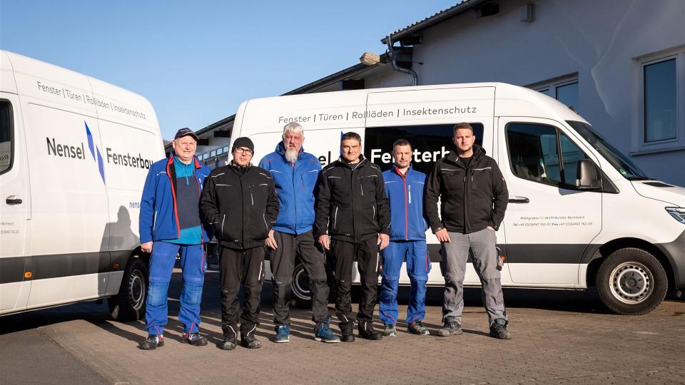 Sechs Mitarbeiter von Nensel Fensterbau stehen nebeneinander in Arbeitskleidung vor einem Firmenfahrzeug mit dem Aufdruck „Fenster | Türen | Rollläden | Insektenschutz“. 