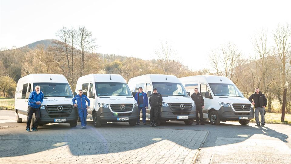 Mehrere Mitarbeiter von Nensel Fensterbau stehen in Arbeitskleidung vor einer Reihe von Firmenfahrzeugen (weiße Mercedes-Transporter) auf einem Betriebshof bei sonnigem Wetter.