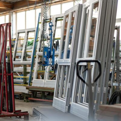 Das Bild zeigt eine Fensterproduktion mit Kunststoffrahmen, die aufrecht in Gestellen lagern. Eine blaue Vakuumhebetechnik hängt von der Decke und dient zum Handling der Fenster.