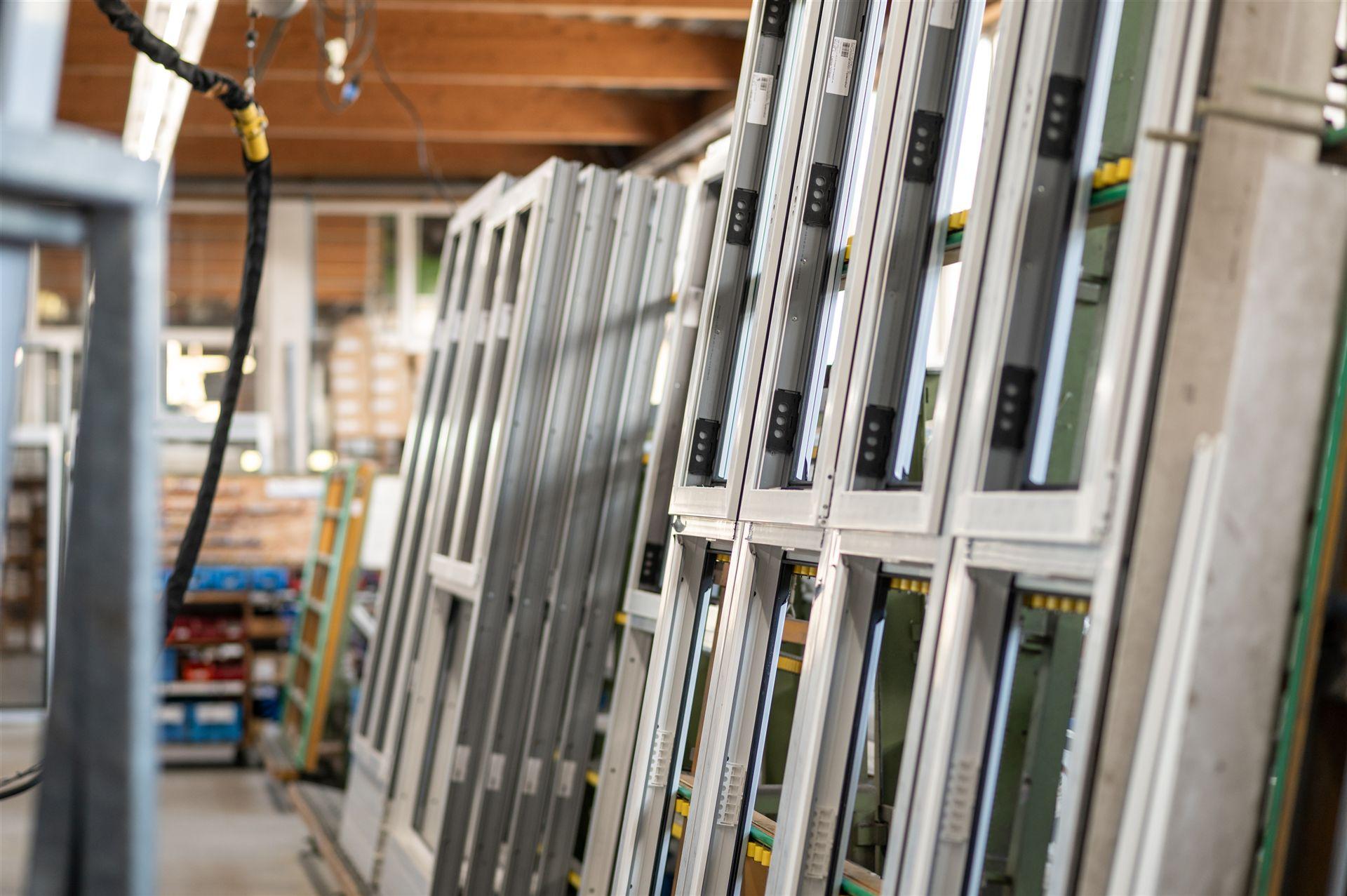 Das Bild zeigt eine Produktionshalle mit mehreren fertiggestellten Fensterrahmen, die ordentlich in Reihen gelagert sind. Im Hintergrund sind Regale, Werkzeuge und Maschinen zu sehen.