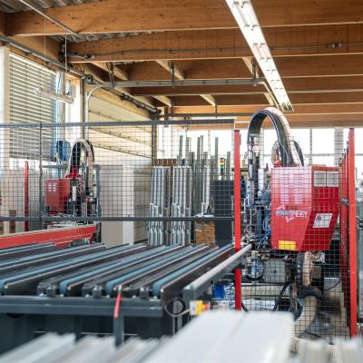 Das Bild zeigt eine Produktionshalle mit einer modernen CNC-Maschine zur Bearbeitung von Profilen für Fenster- oder Türrahmen. Im Vordergrund liegen mehrere Profilstäbe auf einem Fördertisch.