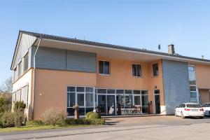 Das Bild zeigt ein modernes, zweigeschossiges Gebäude mit orangefarbener Putzfassade, großen Fenstern im Erdgeschoss und grauen Fassadenelementen im Obergeschoss, vor dem zwei Autos parken.