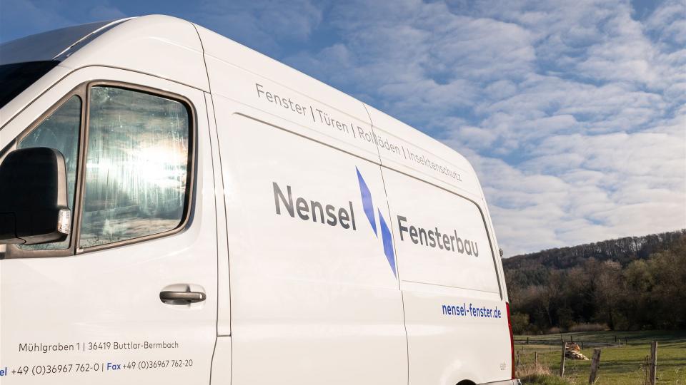 Seitenansicht eines weißen Firmenfahrzeugs mit der Aufschrift „Nensel Fensterbau“ und den Leistungen: Fenster, Türen, Rollläden, Insektenschutz.