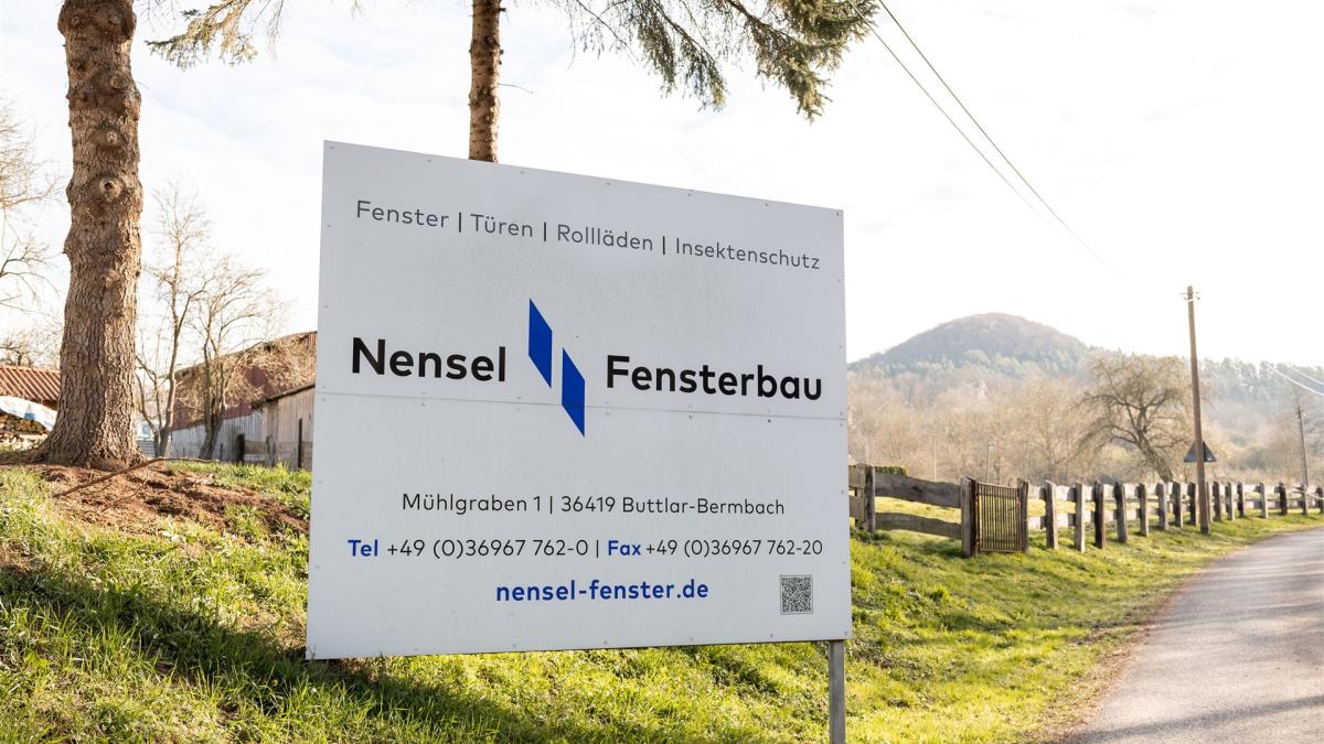 Firmenschild von „Nensel Fensterbau“ mit Kontaktdaten und Leistungen (Fenster, Türen, Rollläden, Insektenschutz) am Straßenrand vor ländlicher Kulisse.
