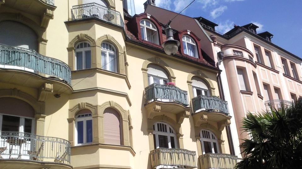 Das Bild zeigt eine Altbaufassade mit dekorativen Fensterrahmen, Rundbogenfenstern, Balkonen mit schmiedeeisernen Geländern und Rollläden.