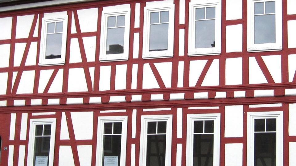 Das Bild zeigt die Fassade eines Fachwerkhauses mit weiß-rotem Holzgebälk und modernen, weißen Sprossenfenstern.