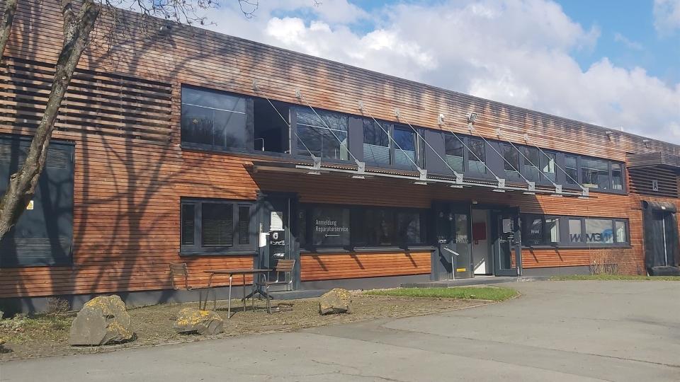 Das Bild zeigt ein modernes Gebäude mit Holzfassade, großen Fenstern und einer Glasüberdachung über dem Eingang. Auf dem Schild steht „Anmeldung Reparaturservice“.
