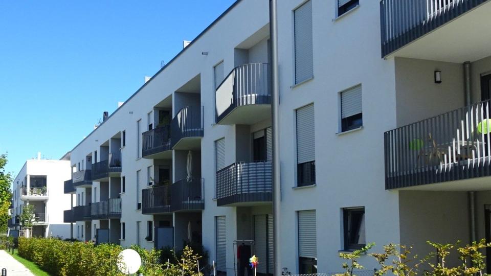 Das Bild zeigt ein modernes Mehrfamilienhaus mit weißen Fassaden, grauen Balkonen und geschlossenen Rollläden unter blauem Himmel.