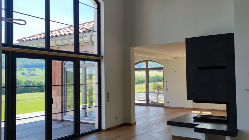 Heller, moderner Wohnraum mit hohen Decken, großen Fenstern, Holzboden und wandintegriertem Kamin. Blick ins Grüne.