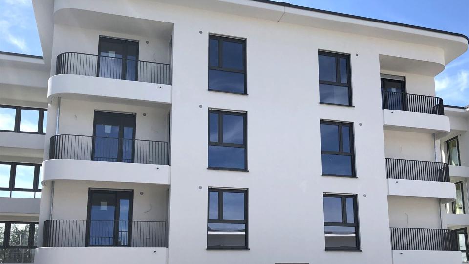Das Bild zeigt ein neu erbautes, modernes Wohngebäude mit weißer Fassade, großen dunklen Fenstern und Balkonen mit schlichten Metallgeländern.