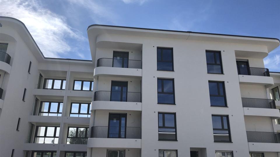 Das Bild zeigt ein modernes, neu gebautes Wohnhaus mit weißer Putzfassade, dunklen Fenstern, abgerundeten Balkonen und klarer Architektur unter blauem Himmel.