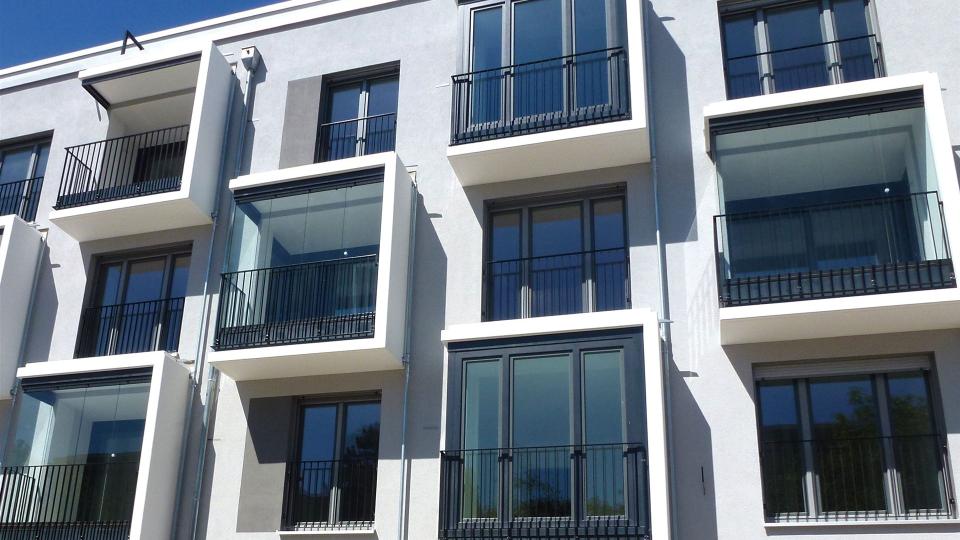 Das Bild zeigt die Fassade eines modernen Wohngebäudes mit bodentiefen Fenstern, französischen Balkonen und teils verglasten Erkern unter blauem Himmel.