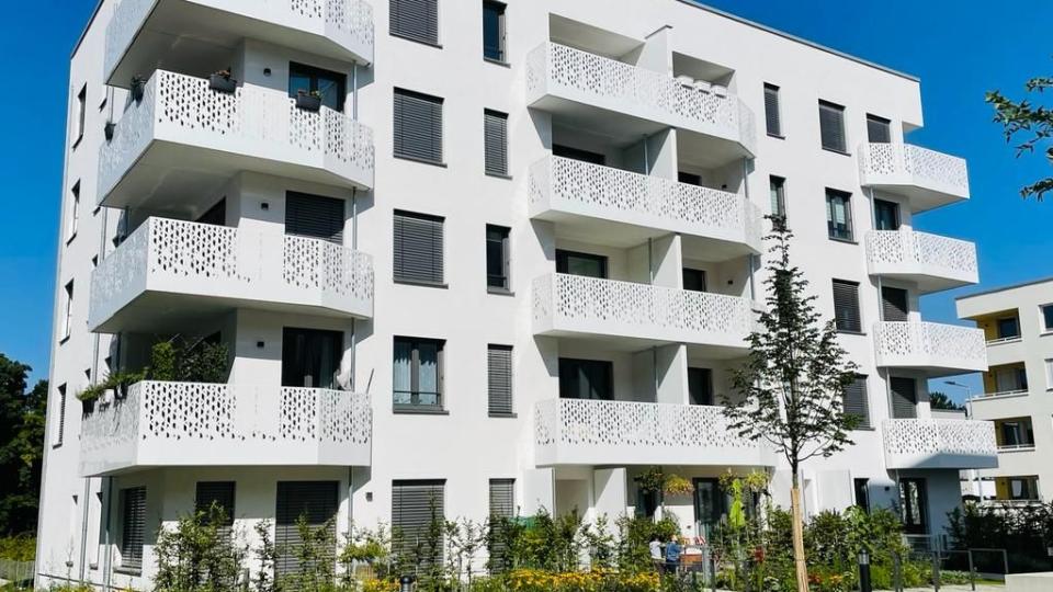 Das Bild zeigt ein modernes Wohnhaus mit weißen Fassaden, dekorativen Balkongeländern und anthrazitfarbenen Fensterläden, umgeben von gepflegter Grünanlage.