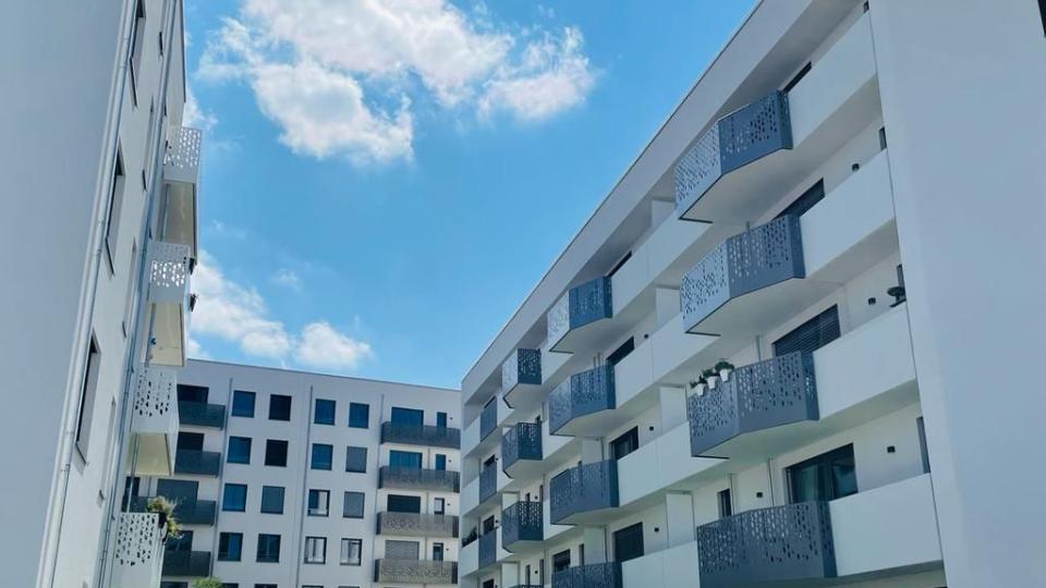 Das Bild zeigt eine moderne Wohnanlage mit weißen Fassaden und dunkelgrauen, gemusterten Balkongeländern. Im Vordergrund befindet sich eine begrünte Hofanlage unter blauem Himmel mit wenigen Wolken.