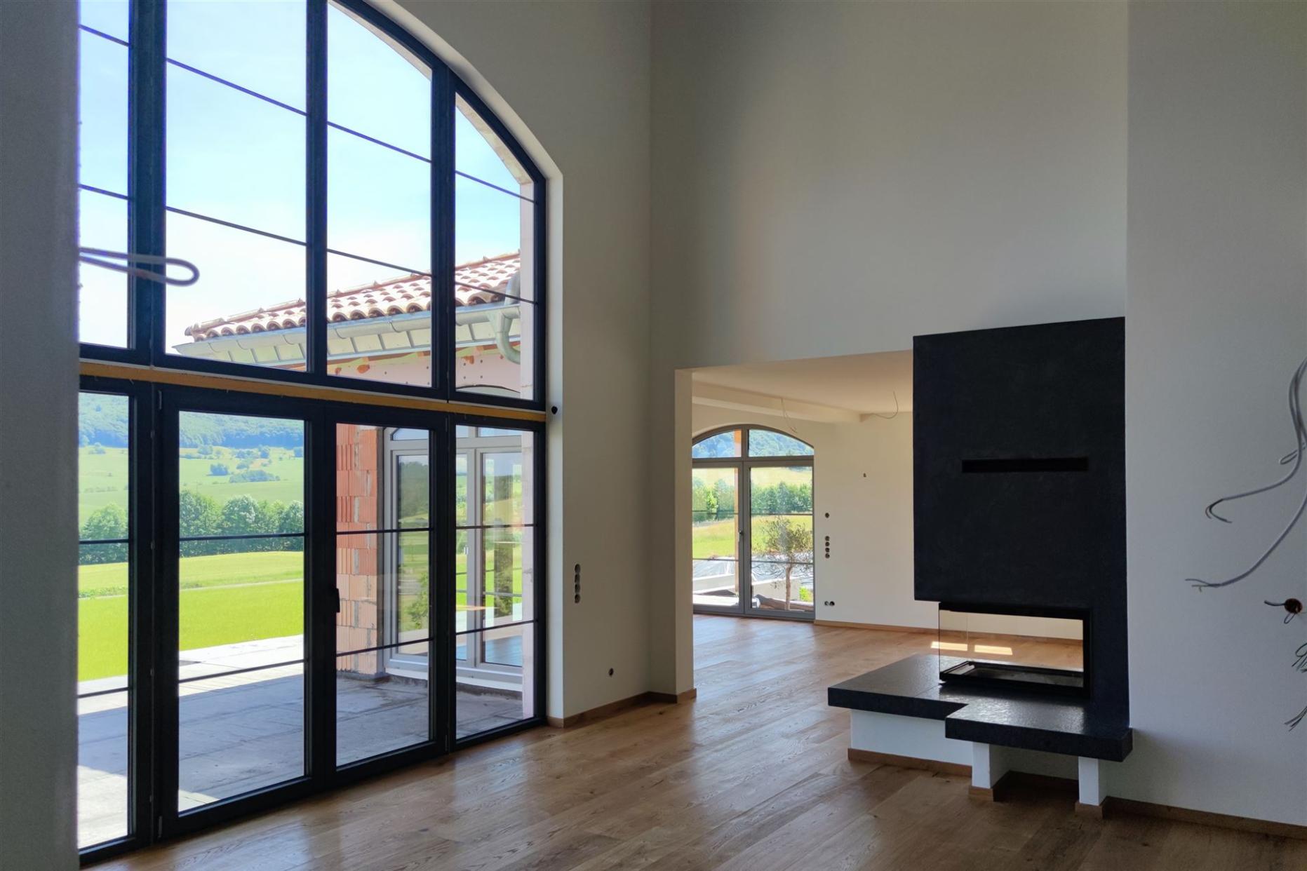 Heller, moderner Wohnraum mit hohen Decken, großen Fenstern, Holzboden und wandintegriertem Kamin. Blick ins Grüne.
