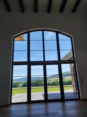 Das Bild zeigt eine große, bogenförmige Fensterfront mit Blick auf eine grüne Landschaft und Hügel unter blauem Himmel.